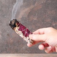 Hand holding a lit Sage and Rose Herb Bundle, with one end smoldering, and a trickle of smoke rising upward from the end.
