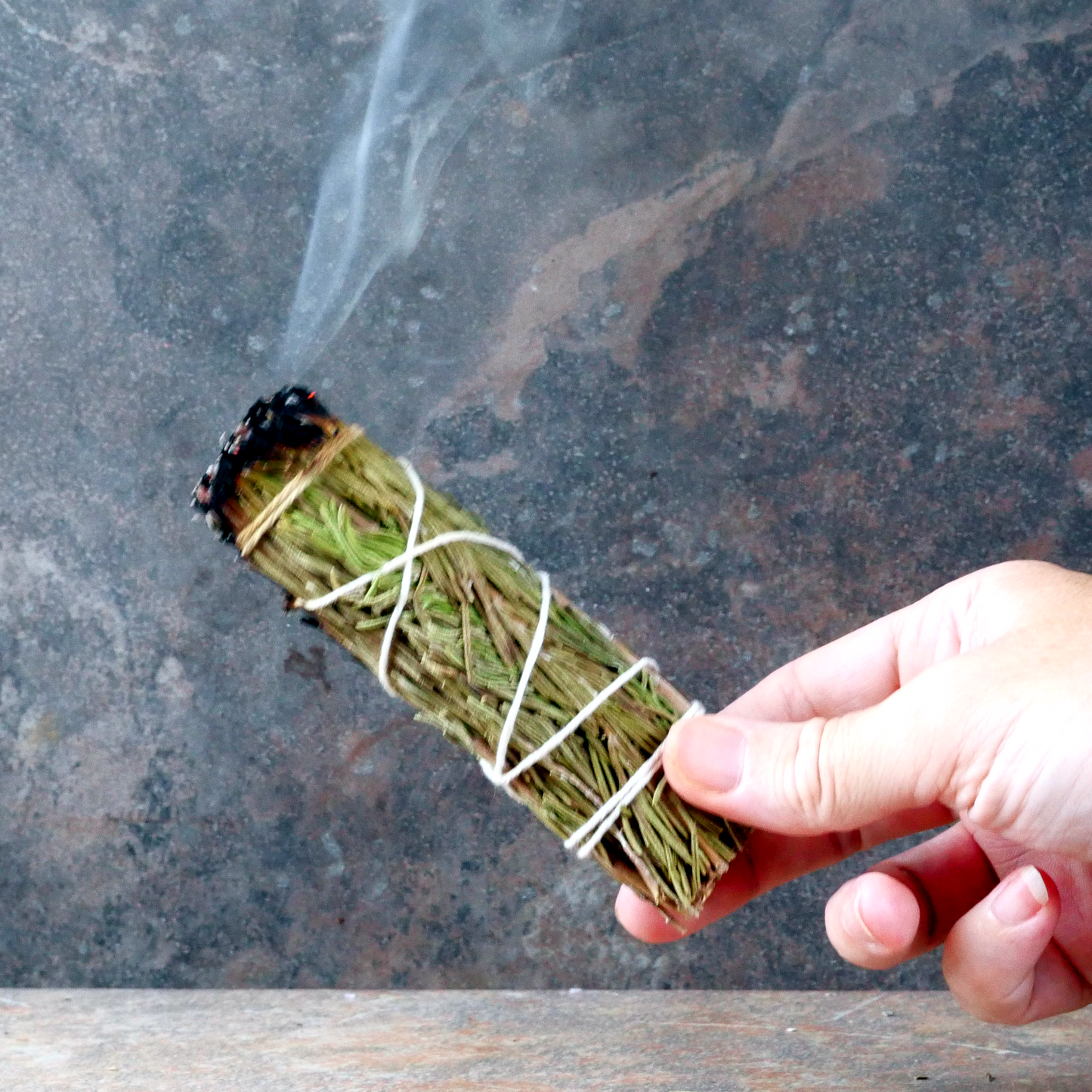 Hand holding a lit Rosemary Herb Bundle, with one end smoldering, and a trickle of smoke rising upward from the end.