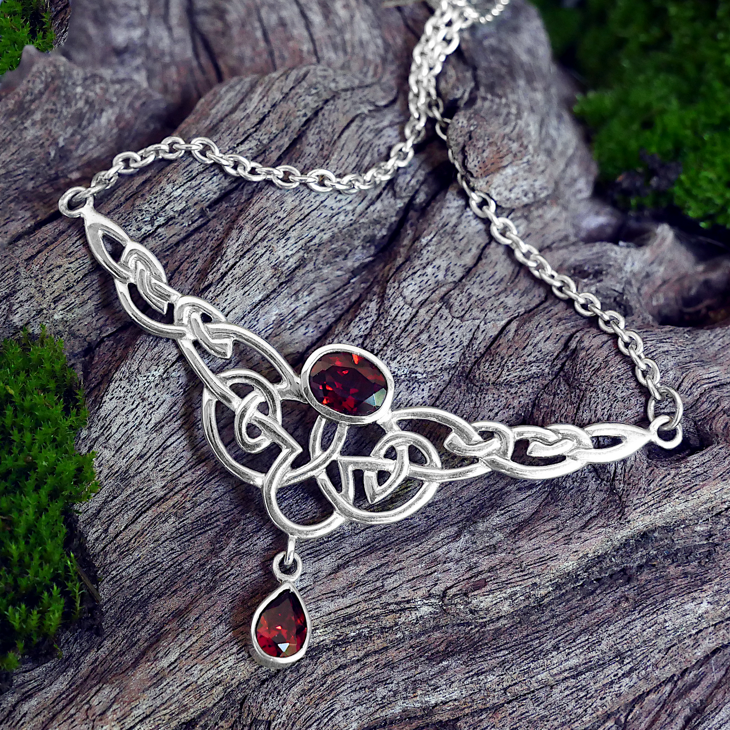Silver Celtic Passion Necklace with garnet gems, delicately displayed on a piece of mossy driftwood for a natural aesthetic.