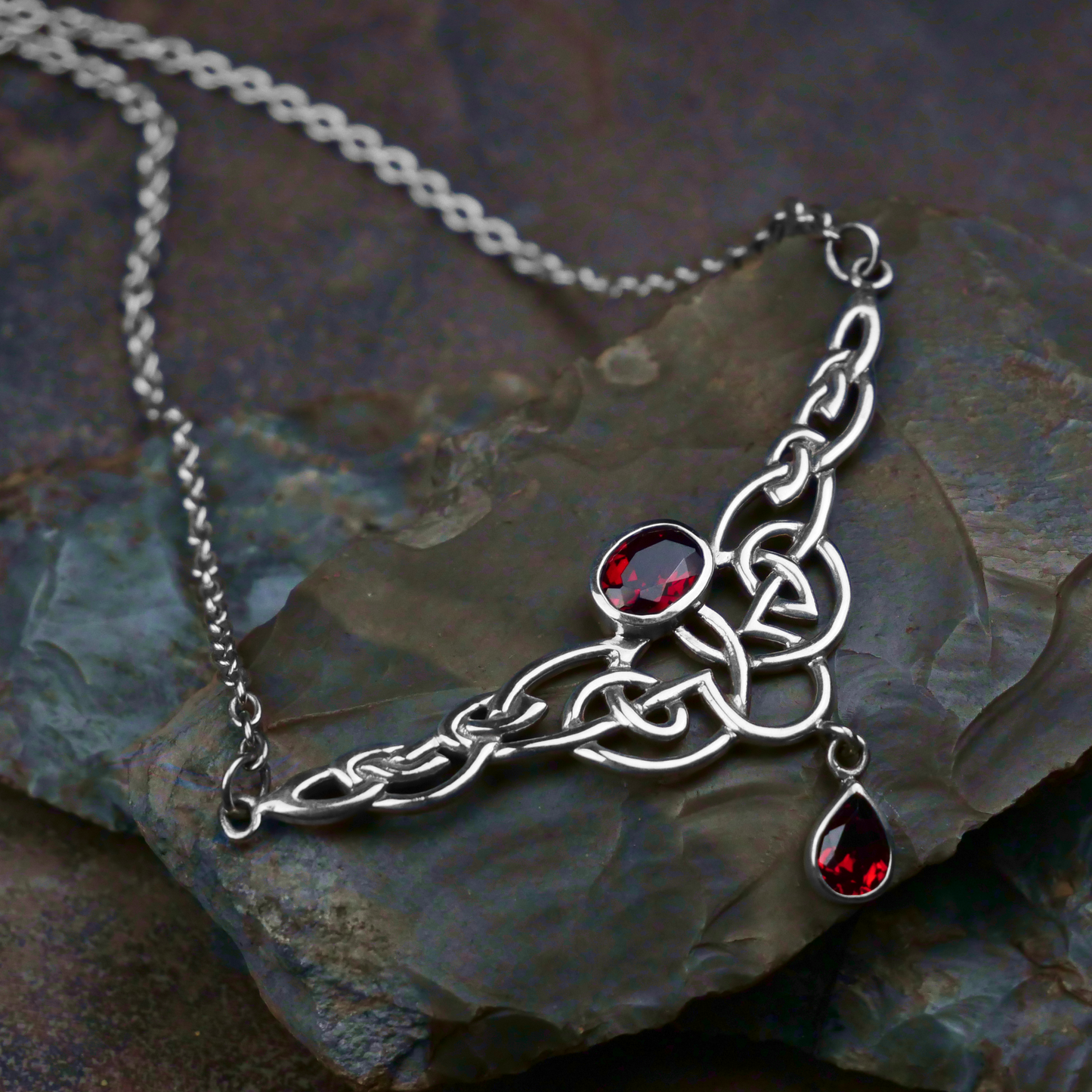 Sterling Silver Celtic Passion Necklace with Celtic knotwork and genuine garnet accents, displayed on a rustic stone surface.