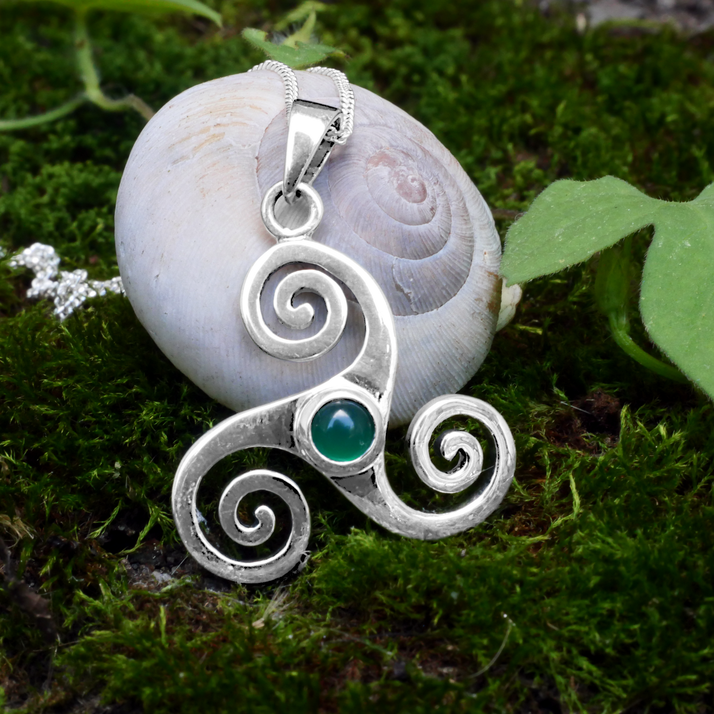Sterling Silver Triskele Pendant with green agate, resting on moss beside a pale white snail shell, mimicing its spiral design.