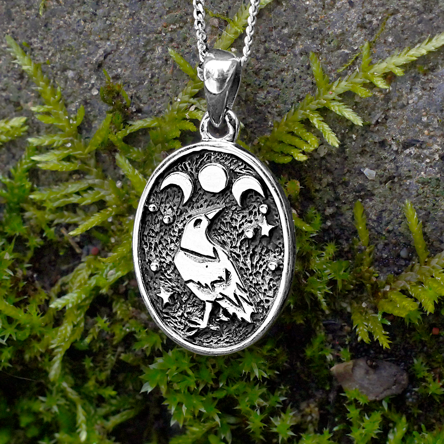 Close-up of the Silver Raven Moon Pendant, featuring a magical raven and triple moon design, displayed on a mossy surface.