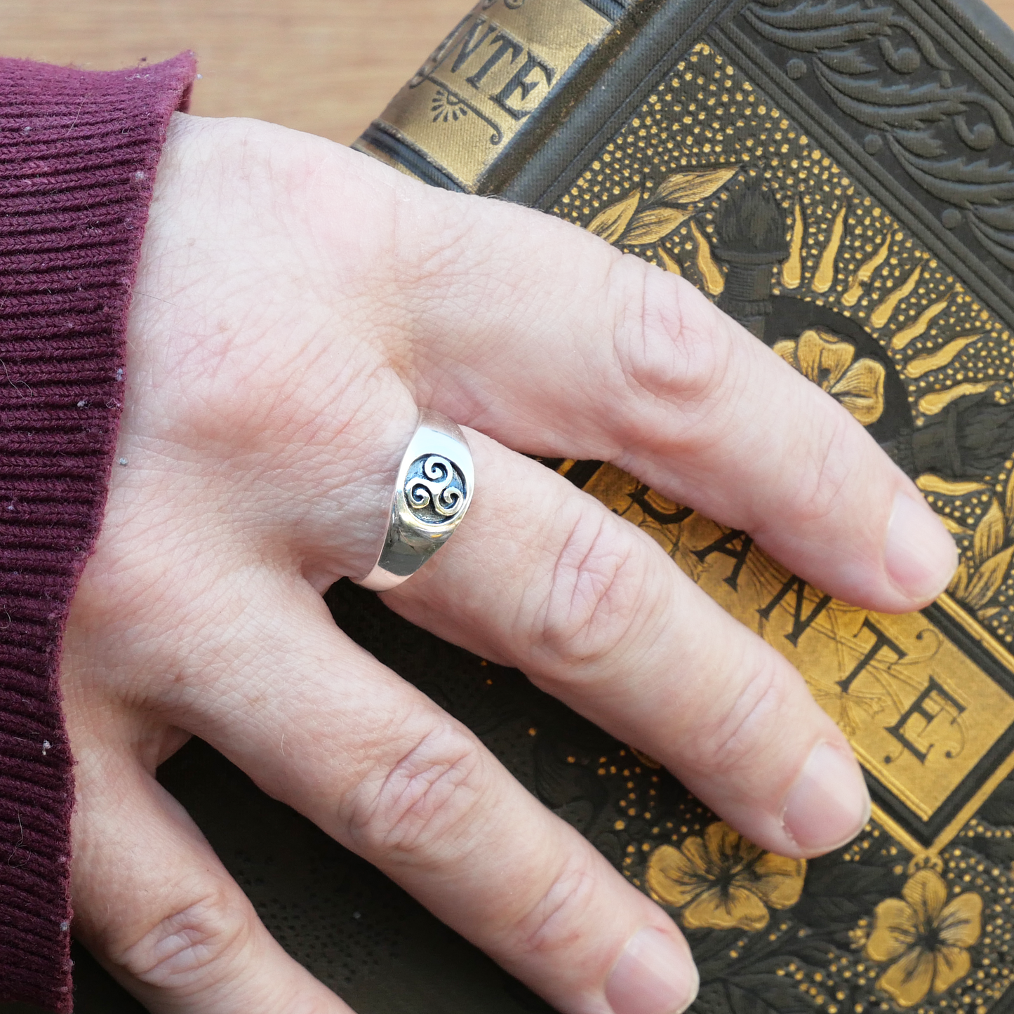Sterling Silver Triskele Signet Ring accented with a Celtic triple spiral design, worn on a hand holding a vintage book.