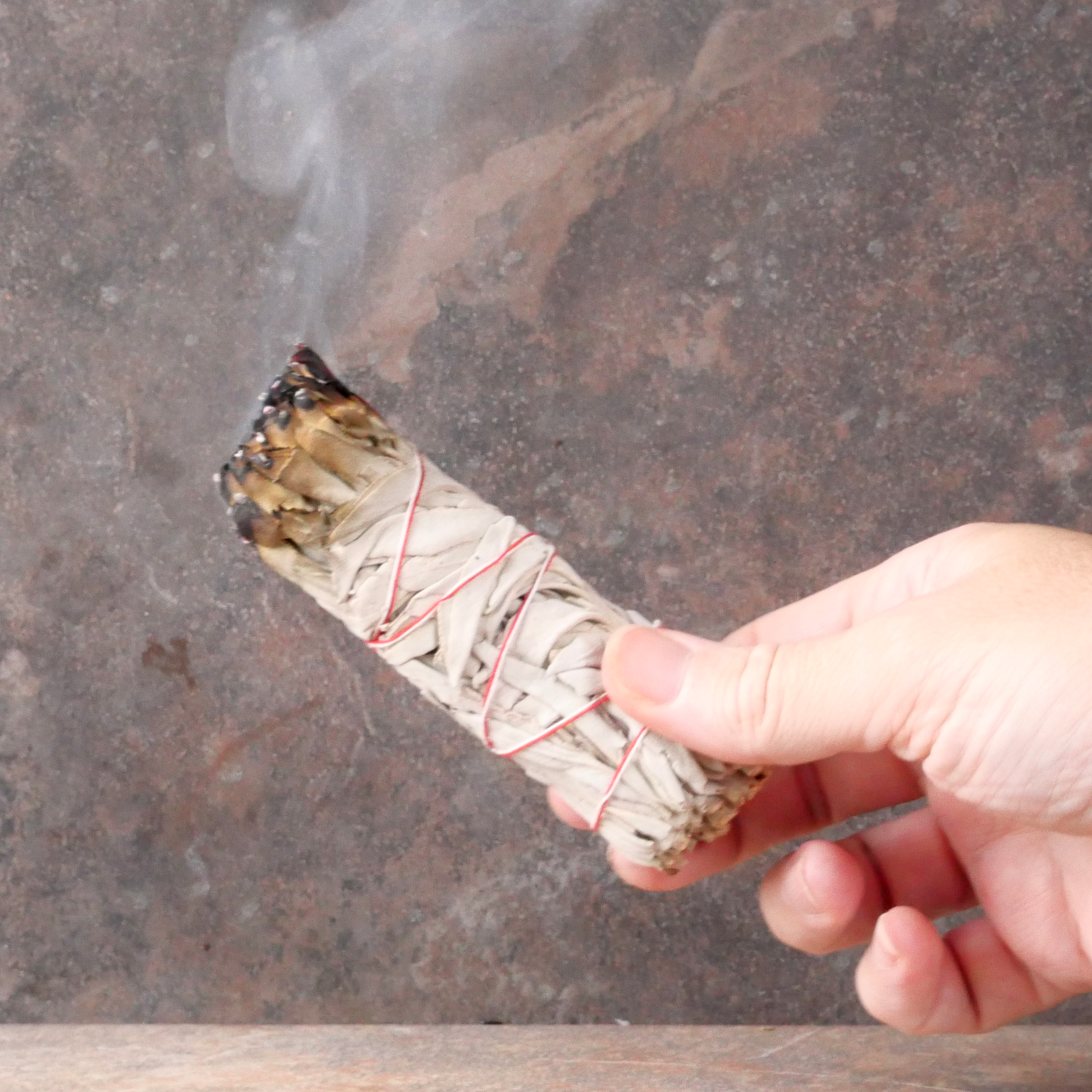 Hand holding a lit White Sage Herb Bundle, with one end smoldering, and a trickle of smoke rising upward from the end.