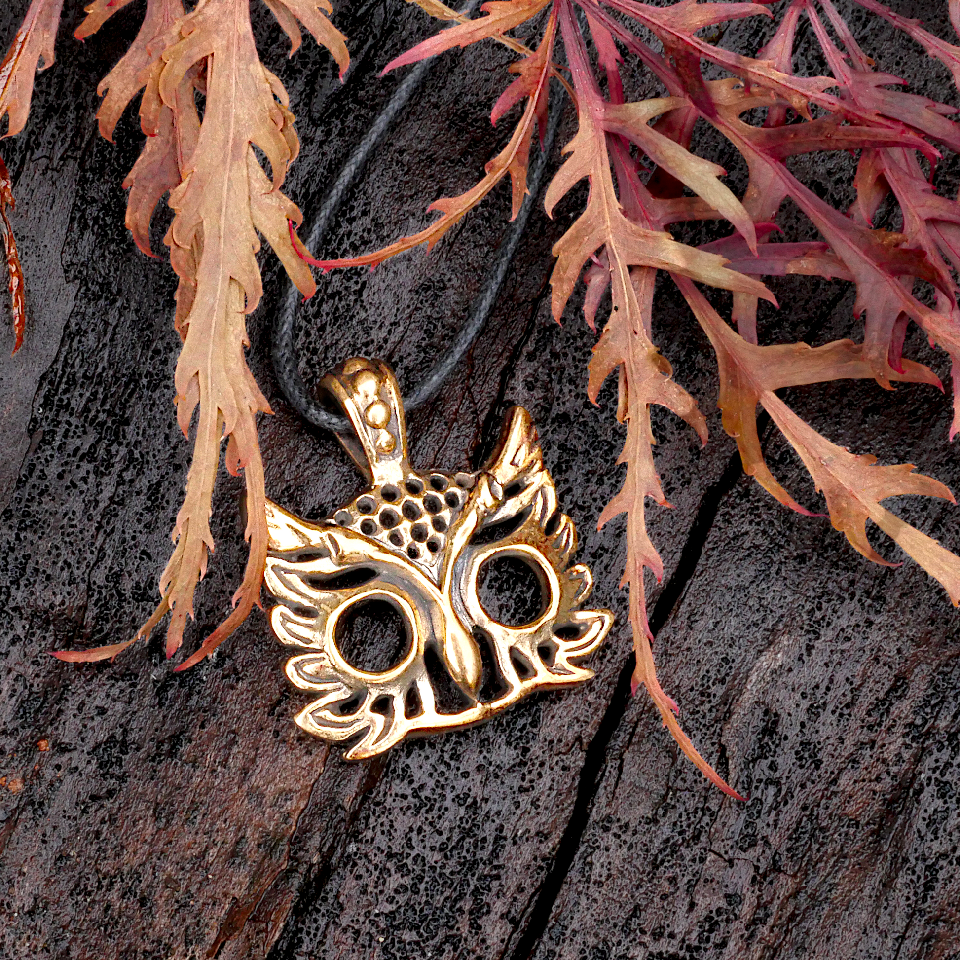 Bronze owl head pendant with large, open eyes, on a dark wood background, framed by delicate red and orange maple leaves.