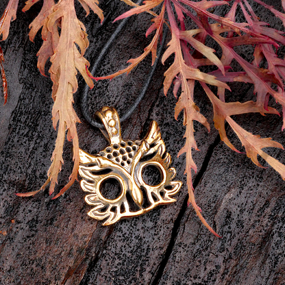 Bronze owl head pendant with large, open eyes, on a dark wood background, framed by delicate red and orange maple leaves.