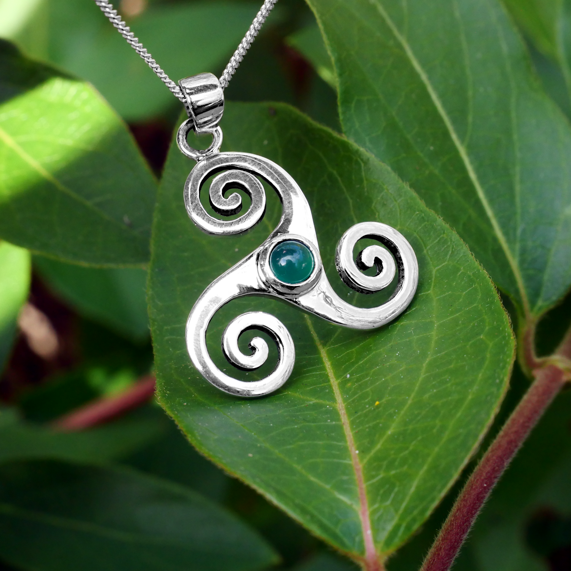 Sterling Silver Triskele Pendant with green agate stone, resting on vibrant green leaves, showcasing its Celtic-inspired design.