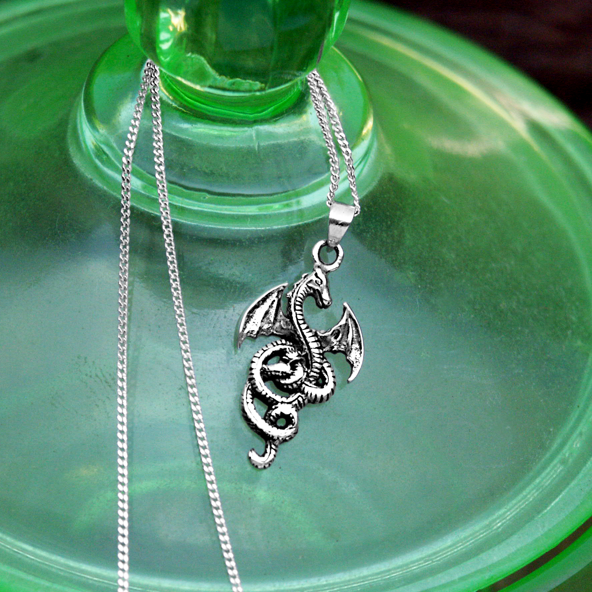 Sterling Silver Coiled Dragon Pendant resting on a surface of luminous green glass, with its chain looped gracefully nearby.