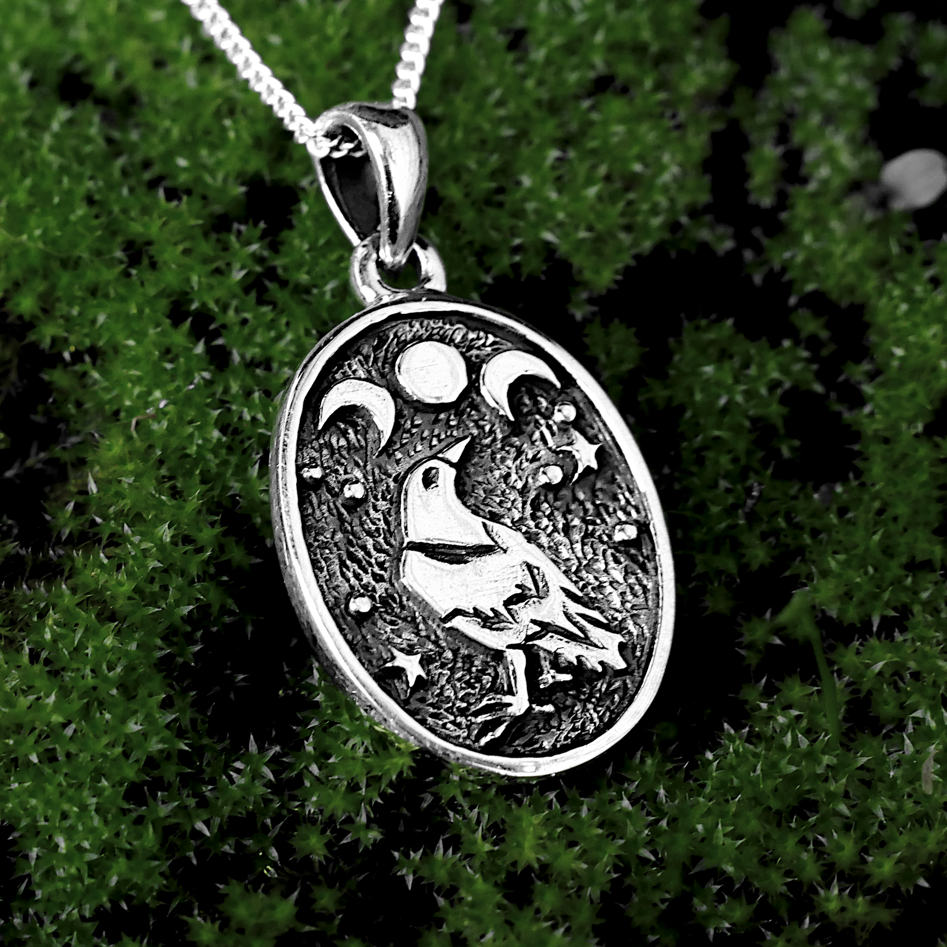 Silver Raven Moon Pendant displayed on a mossy background, emphasizing its nature-inspired design and gleaming highlights.