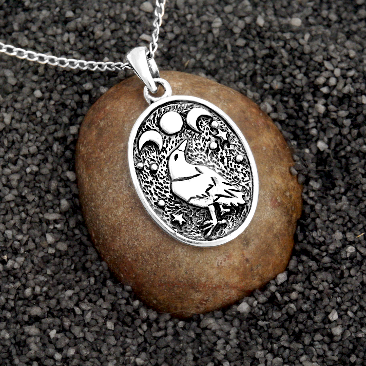 Silver Raven Moon Pendant displayed on a stone and sand background, emphasizing the mystical moon phases and raven design.