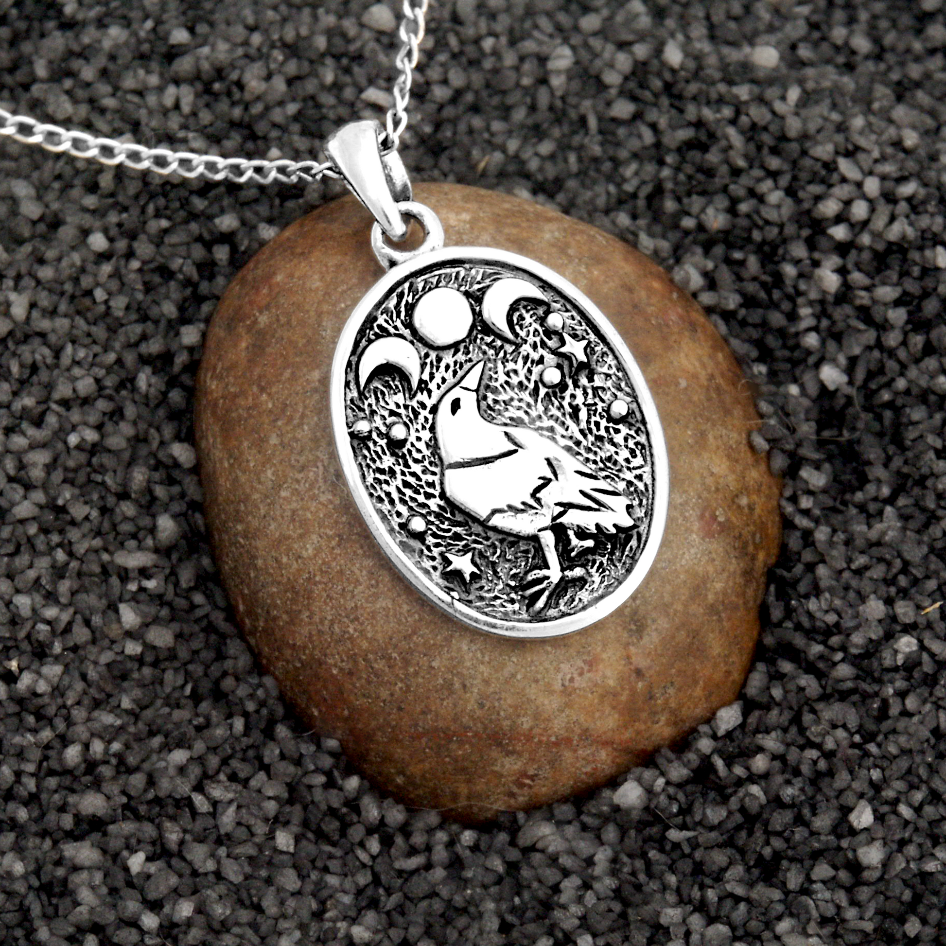 Silver Raven Moon Pendant displayed on a stone and sand background, emphasizing the mystical moon phases and raven design.