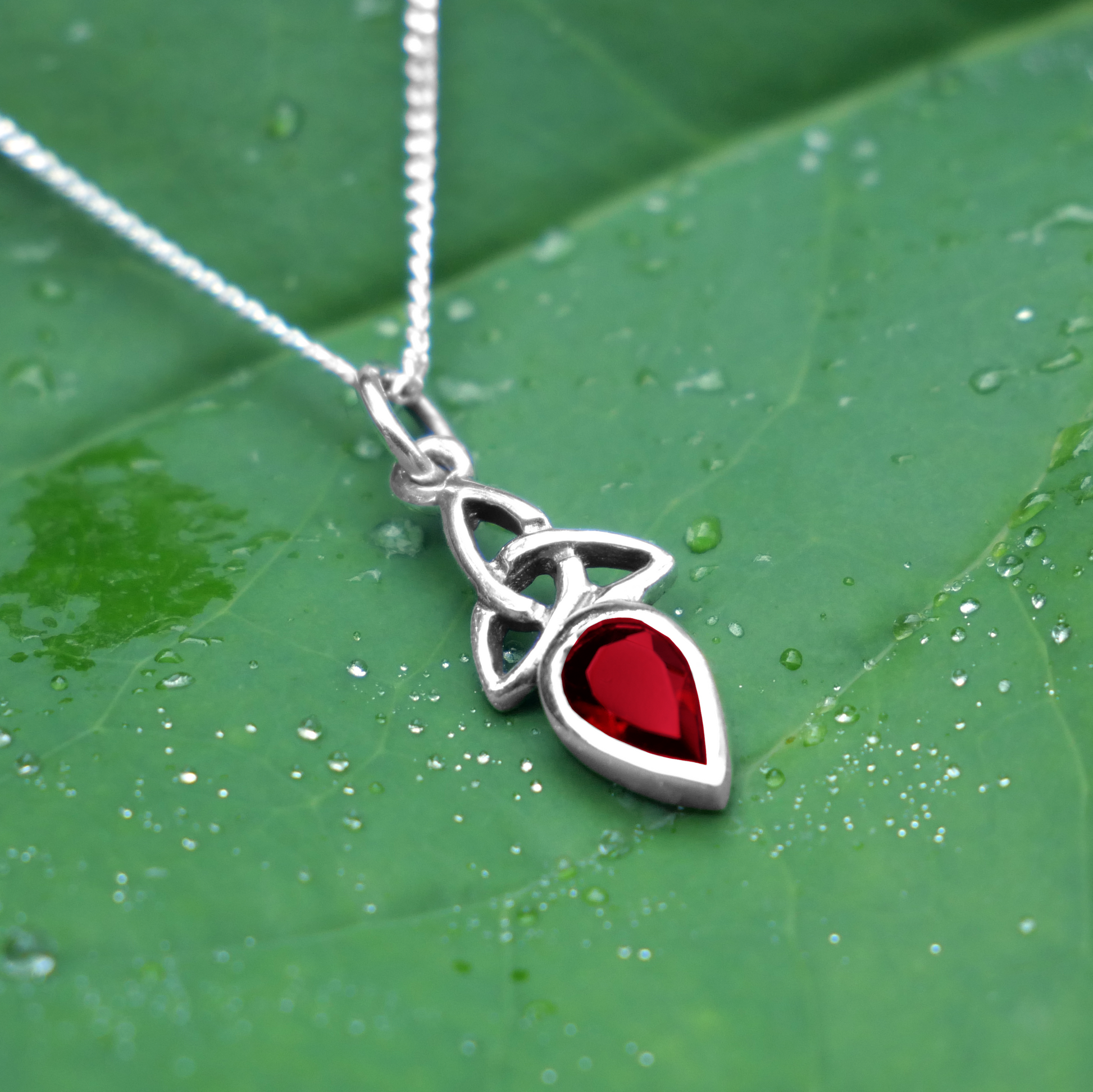 Silver Garnet Trinity Knot Pendant shown on a dewy green leaf, highlighting its garnet gemstone and elegant Celtic knotwork.