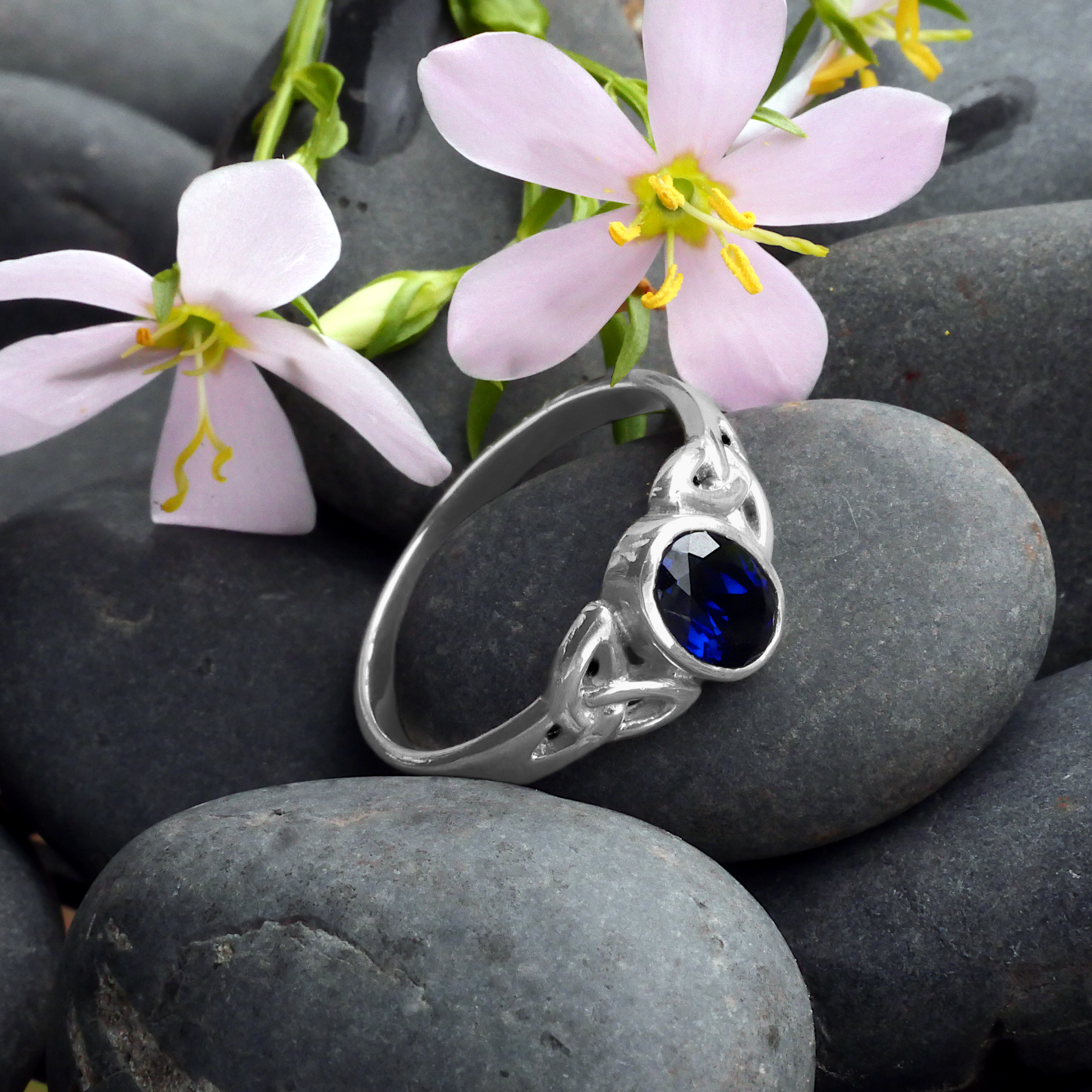 Sterling Silver Sapphire Celtic Ring, resting on smooth black stones with delicate pink flowers in the background.