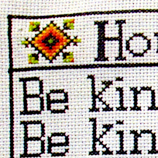 Close-up of one corner of the stitched House Rules cross stitch design, showing black lettering and a small geometric motif.