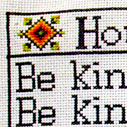 Close-up of one corner of the stitched House Rules cross stitch design, showing black lettering and a small geometric motif.