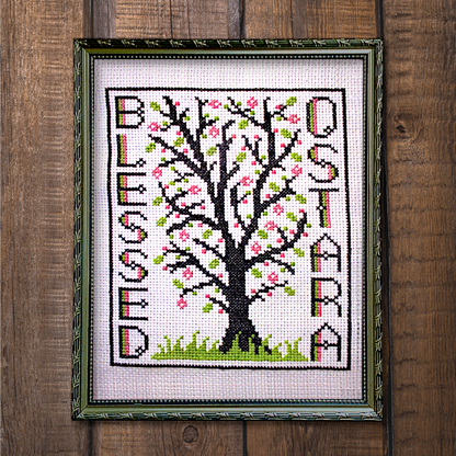 Framed Blessed Ostara cross stitch project, fully stitched and displayed on a wooden background, featuring a tree in bloom.
